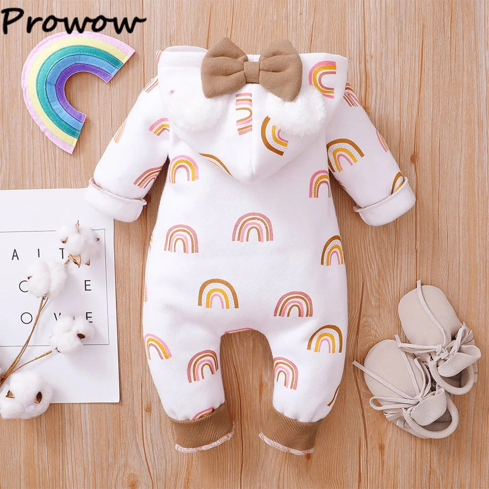 Une combinaison pour bébé avec un motif arc-en-ciel coloré est disposée sur une surface en bois. La combinaison comporte une capuche avec un nœud marron et des pompons moelleux, ressemblant à une Combinaison pour Nouveau-nés. À côté se trouvent des chaussures de bébé beiges, un arc-en-ciel découpé en papier, des fleurs en coton et une étiquette « BABY PREMA ».
