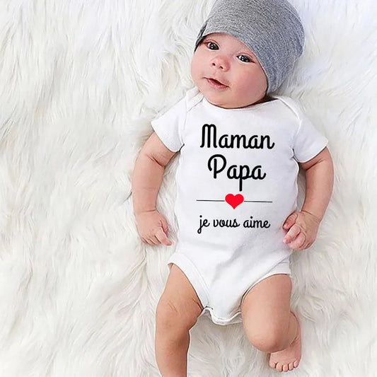 Un bébé portant un bonnet gris et une combinaison blanche est allongé sur une couverture douce et blanche en fourrure. La combinaison porte le texte français « Maman Papa, je vous aime » avec un cœur rouge sous les mots. Parfait comme cadeau bébé pour la fête des mères et pères, le bébé regarde l'appareil photo avec une expression douce. Le produit s'appelle Body Coton Fête des Mères & Pères de BABY-PREMA.