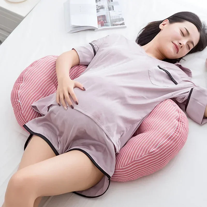 Une femme enceinte est allongée sur un lit, utilisant un Oreiller de Grossesse en Coton rayé en forme de U de BABY PREMA pour se soutenir. Elle porte un pyjama en satin rose clair et semble se reposer paisiblement, les yeux fermés.