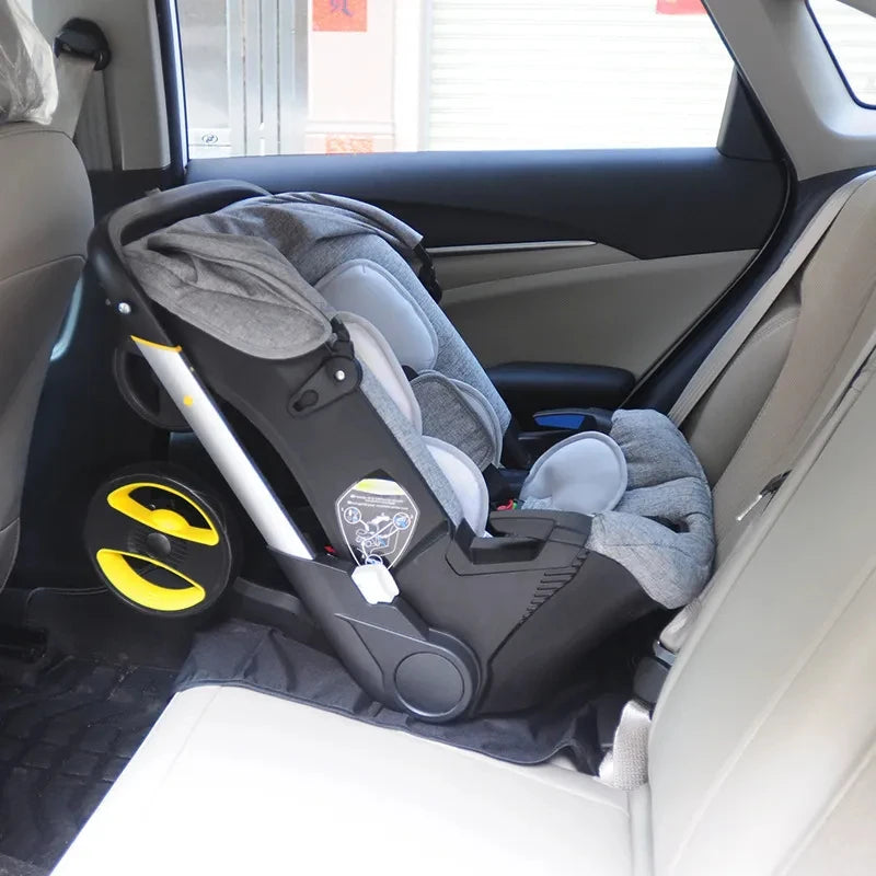 Une Poussette 4 en 1 Cosy Bébé BABY-PREMA grise est installée sur la banquette arrière d'une voiture, face à l'arrière. Le siège auto comporte des côtés rembourrés, un harnais de sécurité et une poignée réglable. Il repose sur un tapis noir protégeant le siège du véhicule.