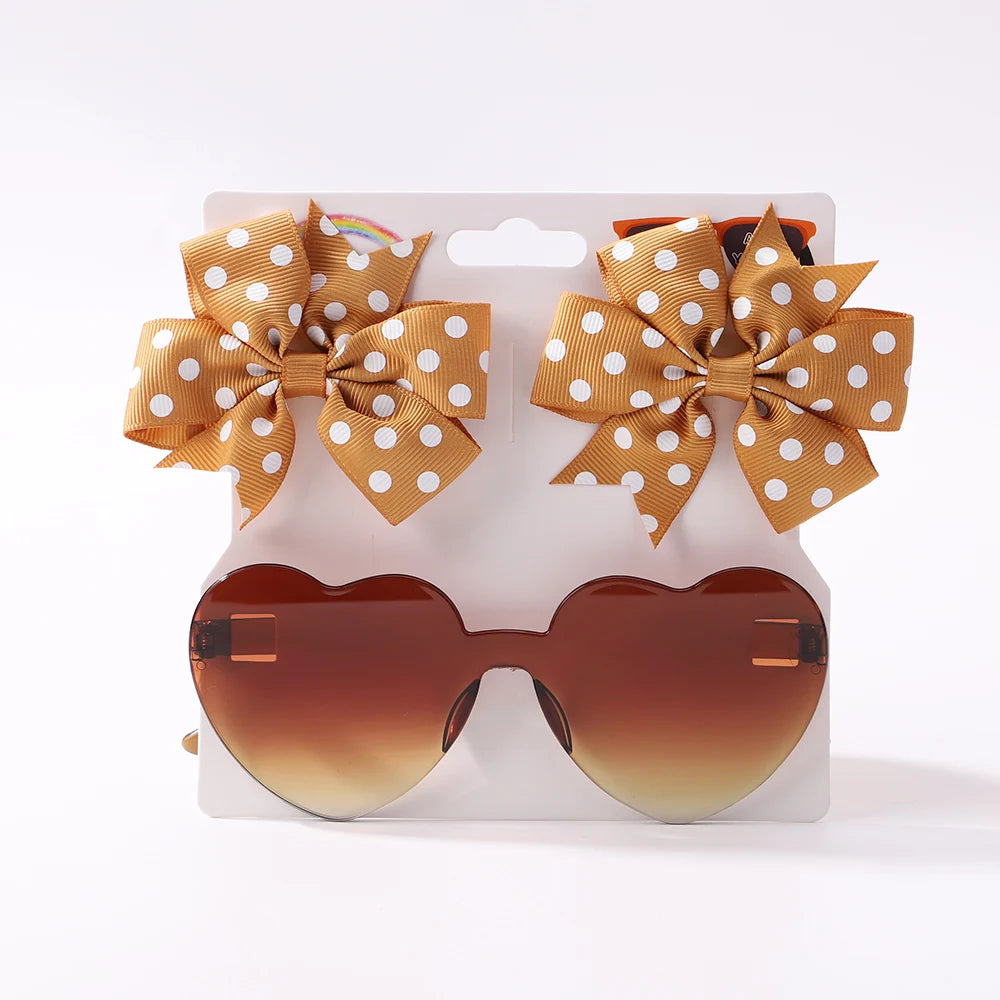 Un ensemble d'accessoires bébé | Les Lunettes de Cheveux pour Bébé de BABY PREMA, composées d'une paire de lunettes de soleil pour bébé en forme de cœur avec des verres marron dégradés, sont présentées sous deux bandeaux à nœuds en nylon assortis de couleur moutarde à pois blancs.