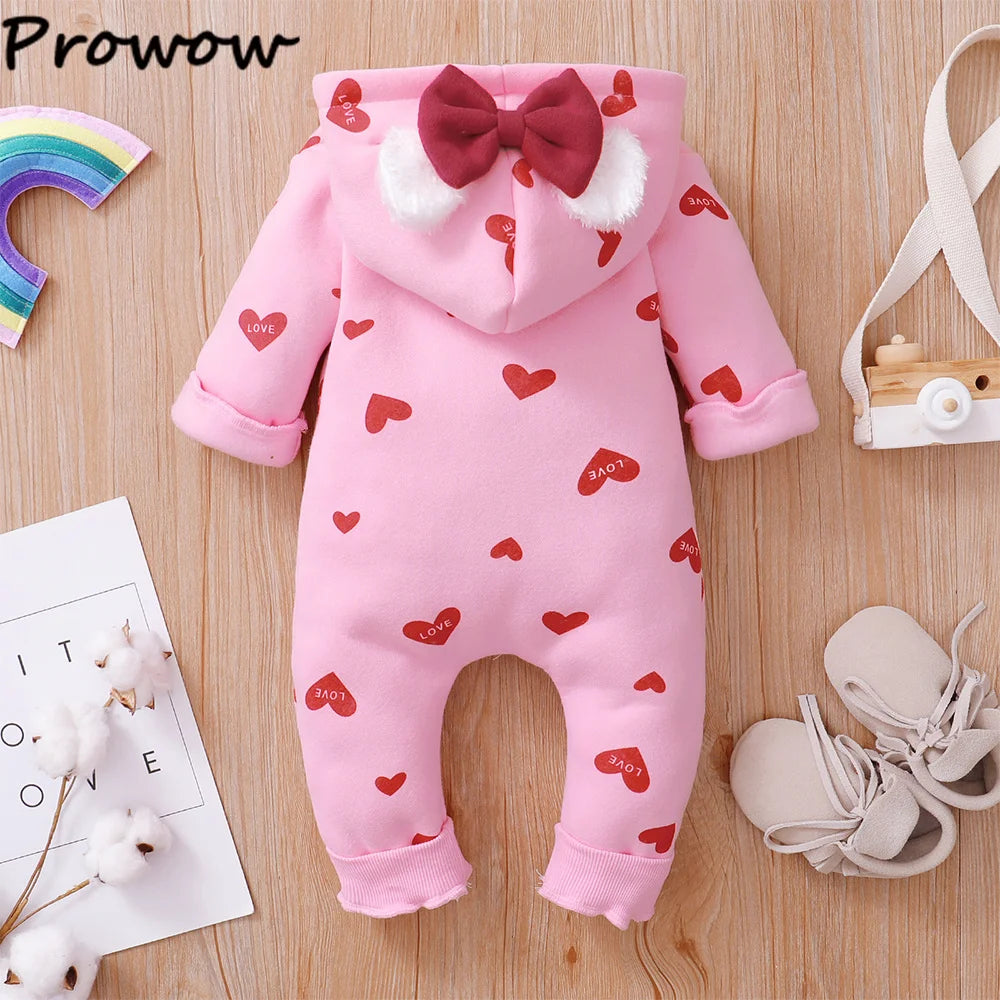 Une Combinaison pour Nouveau-nés BABY PREMA de couleur rose avec une capuche, avec des motifs de cœurs rouges avec le mot « Love » écrit à l'intérieur. La capuche est ornée d'oreilles duveteuses blanches et d'un nœud rouge. La tenue, parfaite pour l'hiver, est posée à plat sur un plan en bois aux côtés de chaussures de bébé et d'une décoration murale en forme d'arc-en-ciel.