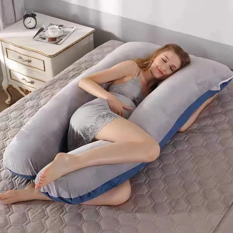 Une femme en tenue de nuit est confortablement allongée sur un lit, serrant dans ses bras un grand Coussin Maternité Multifonction en forme de U de BABY PREMA. Elle sourit les yeux fermés, ce qui suggère qu'elle est détendue et à l'aise. Son lit a une couverture matelassée grise et il y a une table de nuit blanche avec des objets dessus à proximité.
