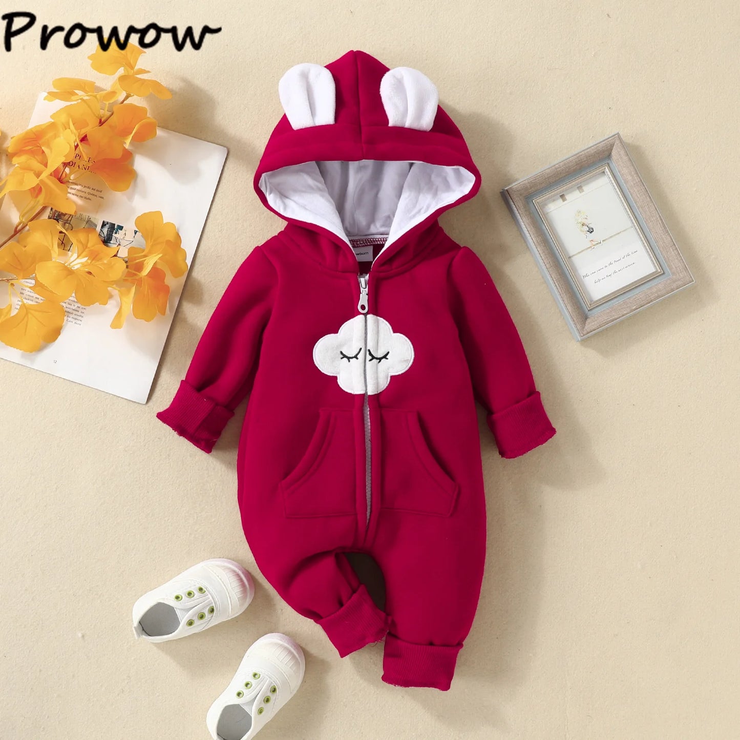 Une Combinaison pour Nouveau-nés BABY PREMA rouge avec une capuche avec des oreilles de lapin blanches et un joli visage blanc avec les yeux fermés sur le devant. La tenue, parfaite comme vêtements d'hiver pour bébé, est disposée sur un fond beige aux côtés d'une paire de chaussures blanches, de feuilles jaunes et d'une photo encadrée.