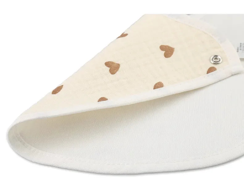 Gros plan d'un bavoir beige avec des motifs de coeurs marron, partiellement plié pour révéler un intérieur blanc et un bouton pression en métal pour la fermeture. Ce style Bandana au design chic, que l'on retrouve dans de nombreuses collections BABY-PREMA Bavoirs Motifs Bébé 2 Pièces, ajoute à la fois fonctionnalité et style à la tenue de votre bébé.