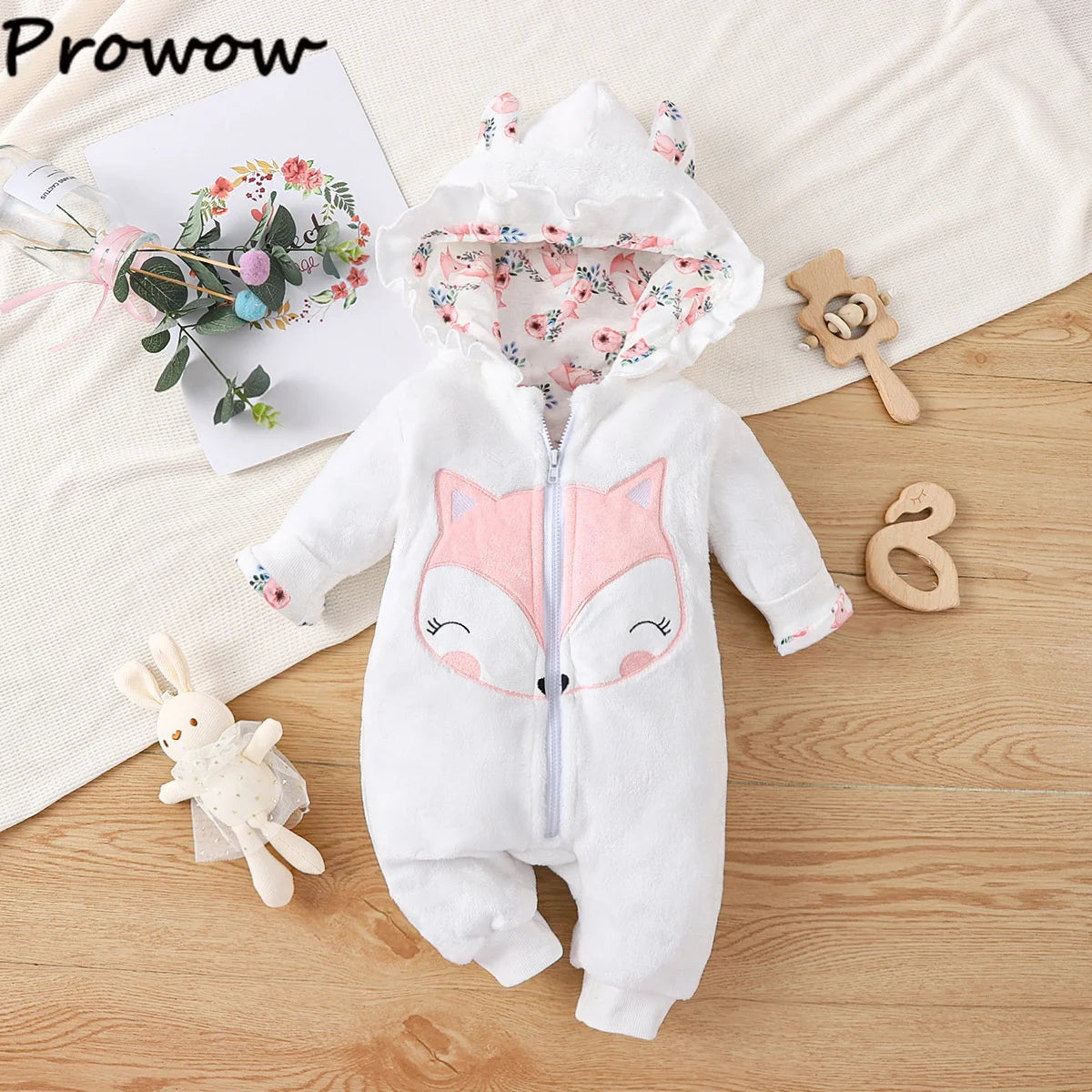Une combinaison pour bébé avec un joli visage de renard sur le devant. La Combinaison pour Nouveau-nés est dotée d'une capuche à oreilles de renard et est disposée sur une surface en bois. Autour de lui se trouvent des jouets pour bébé, un bouquet floral et un chiffon doux en arrière-plan. Le mot « BABY PREMA » se trouve dans le coin supérieur gauche.