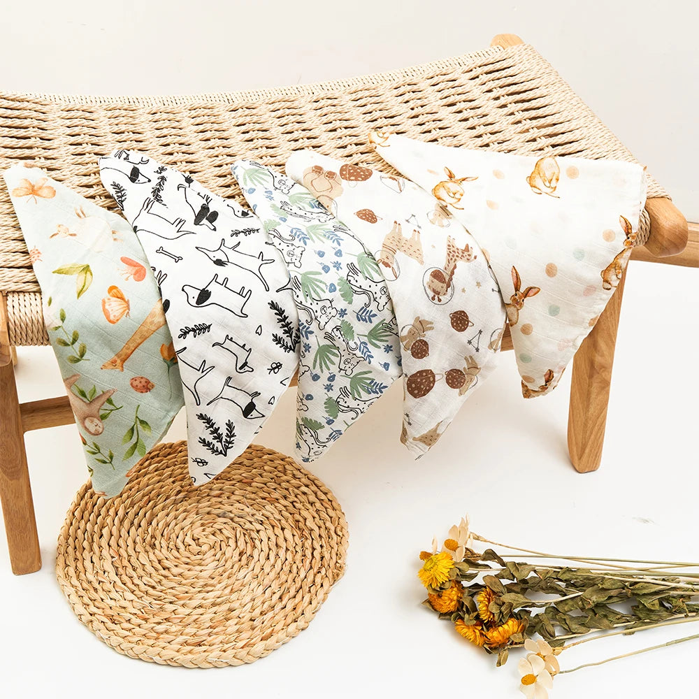 Une collection de serviettes en tissu Langes Lot de 5 pièces Mousseline Coton & Bambou drapées sur un banc en osier, avec un set de table tissé naturel et quelques fleurs séchées posées à côté, évoquant une atmosphère chaleureuse et rustique. Par BÉBÉ PREMA.