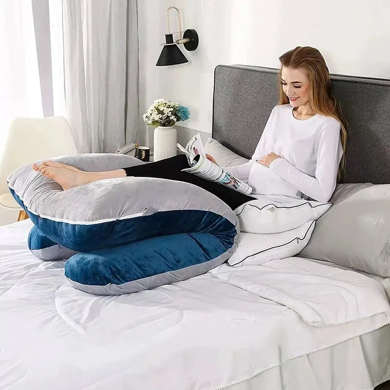 Une femme enceinte est assise sur un lit, confortablement soutenue par un grand Coussin Maternité Multifonction de BABY PREMA. Elle lit un magazine, vêtue d'une chemise blanche à manches longues et d'un pantalon noir. Le lit est soigneusement fait et le fond comprend un décor gris et blanc.