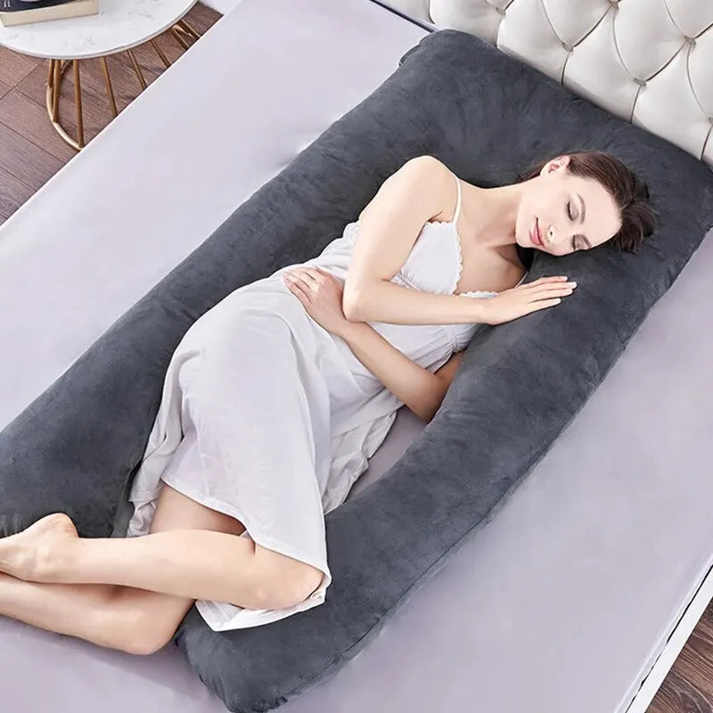 Une femme en chemise de nuit blanche dort sur un lit, serrant dans ses bras un grand Coussin Oreiller Femmes Enceinte de BABY PREMA. Le lit a un drap gris et une tête de lit capitonnée. Il y a une table d'appoint ronde avec un livre et une boisson à côté du lit. La pièce dégage une atmosphère calme et paisible.