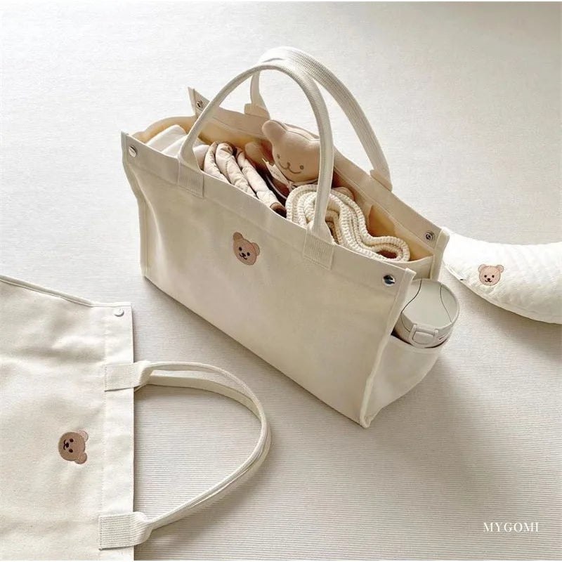 Un sac fourre-tout en toile bien rangé avec un joli emblème en forme de visage d'ours, rempli d'essentiels et de peluches BABY-PREMA Enfant, ainsi qu'un accessoire en tissu assorti.