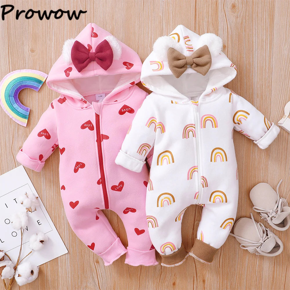 Deux adorables BABY PREMA Barboteuses Combinaison à Capuche sont posées sur une surface en bois. Celui de gauche est rose avec des motifs de cœurs et un nœud rouge ; celui de droite est blanc avec des motifs arc-en-ciel et un nœud marron. A proximité se trouvent des chaussures de bébé, un petit arc-en-ciel au crochet et une carte sur laquelle est écrit « petit amour ».
