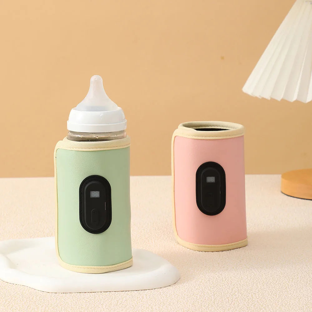 Deux BABY-PREMA Chauffe-Biberon Portable multifonction avec affichage numérique de la température sont enveloppés dans des housses de couleurs pastel, une verte et une rose. Ils sont placés sur une surface beige avec une lampe moderne en arrière-plan. Le décor semble minimaliste avec des tons doux et neutres.