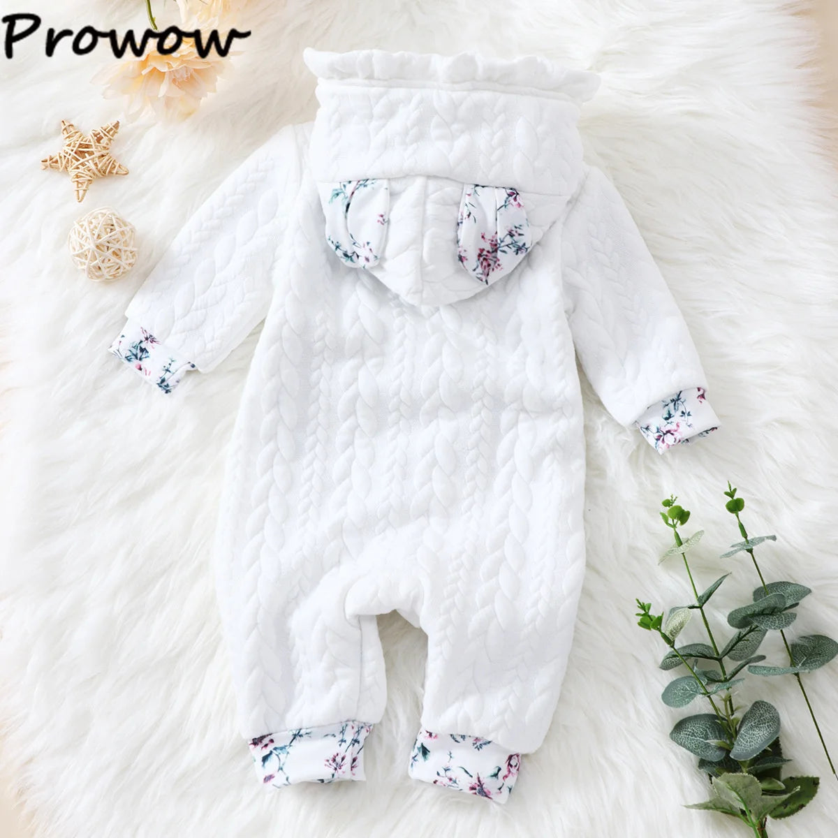 Une combinaison bébé matelassée blanche avec une capuche et des imprimés floraux sur les poignets et à l'intérieur de la capuche. La combinaison pour nouveau-né est disposée sur une douce surface semblable à une fourrure blanche, avec des décorations en forme d'étoile et des feuilles d'eucalyptus vertes à proximité. Le nom de la marque « BABY PREMA » est visible dans le coin supérieur gauche.