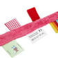Un jouet sensoriel coloré pour bébé comprenant une sangle en peluche rose avec diverses textures et motifs de tissu, un carré rouge, des rubans à carreaux et à rayures et un anneau noir avec des instructions d'entretien attachées - Attache Sucette de LABEL LABEL.