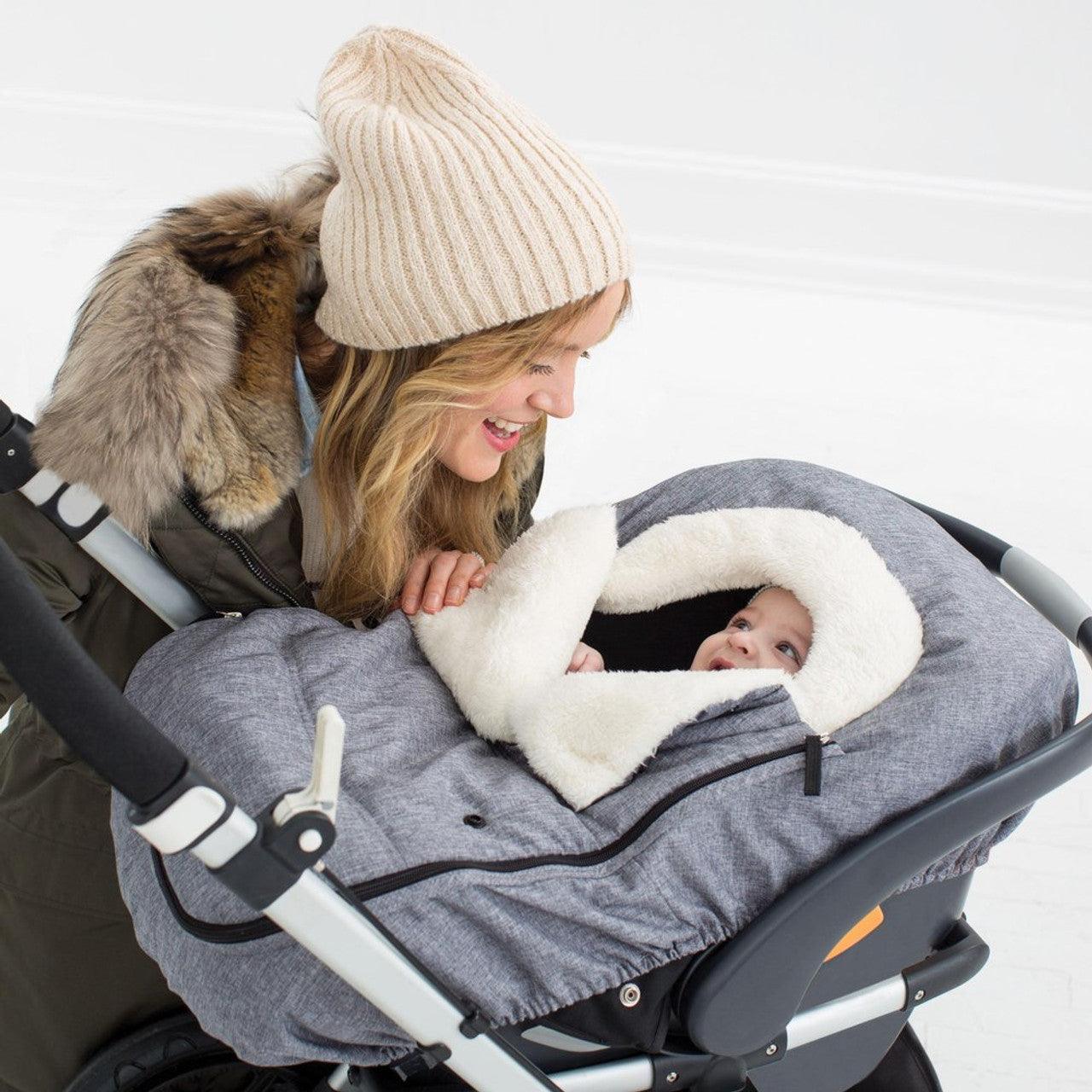 Une femme souriante coiffée d'un chapeau d'hiver regardant affectueusement un bébé, emmitouflé dans une poussette douillette lors d'une journée glaciale, se prépare pour le bain du BABY PREMA Couverture Siège Auto & Poussette Bébé.