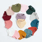 Un assortiment coloré d'accessoires pour bavoirs bébé BABY PREMA disposés en motif circulaire sur un fond blanc.