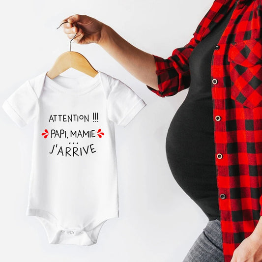 Une personne enceinte vêtue d'une chemise à carreaux rouge tient un bébé blanc sur un cintre en bois. Ce joli **Body Manches Courtes Coton** à manches courtes comporte le texte français qui dit « Attention !!! Papi, Mamie... J'arrive », se traduisant par « Attention !!! Grand-père, Grand-mère... Je' J'arrive." **BÉBÉ-PREMA**