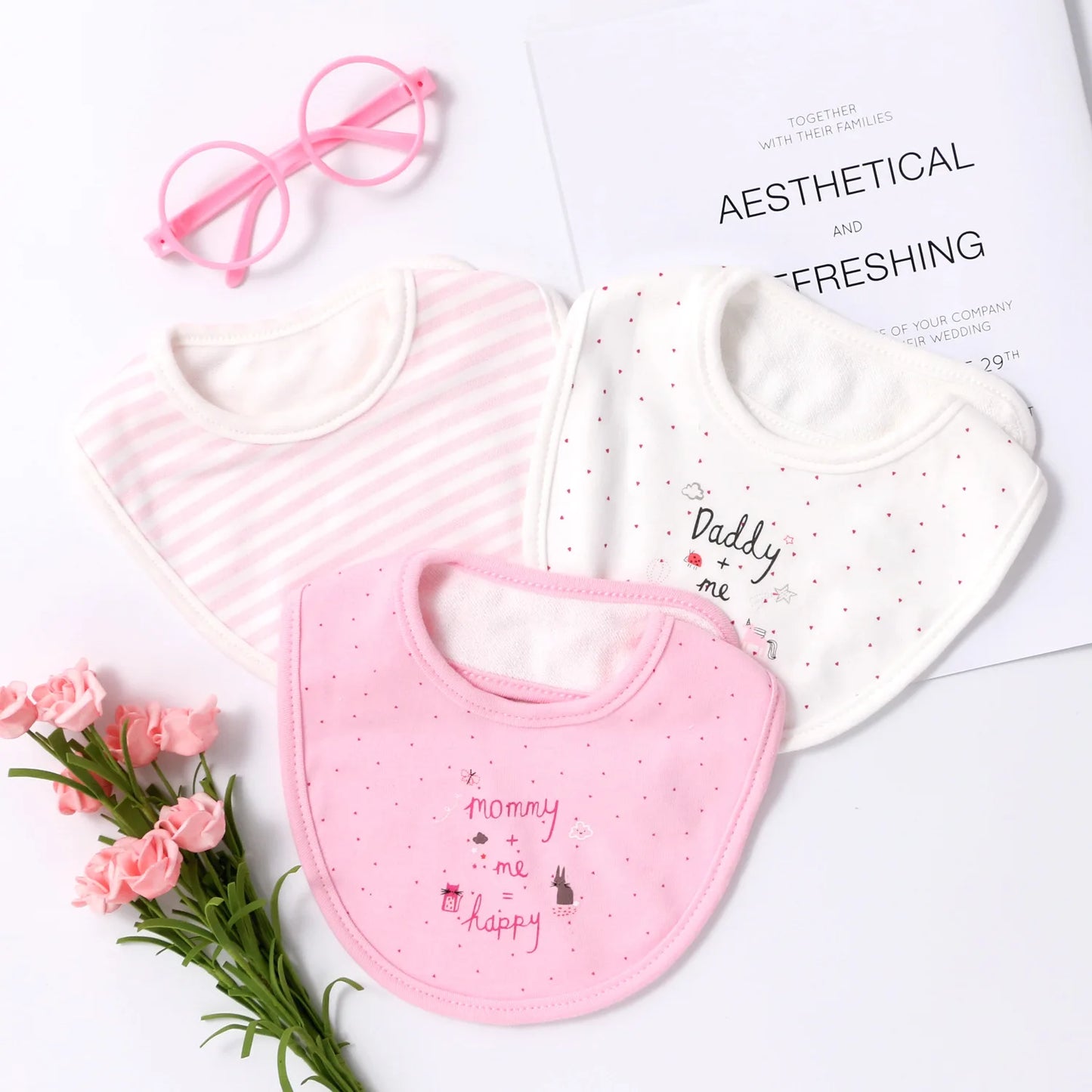 Trois bavoirs pour bébé sont disposés sur un fond blanc, accompagnés d'une paire de lunettes de soleil roses pour bébé et d'un petit bouquet de fleurs roses. Le Lot de 3 pièces Bavoirs Bébé de BABY-PREMA a de jolis designs ; l'un est rose et rayé, l'autre est blanc avec des points et du texte et l'autre est rose avec du texte en pointillés.