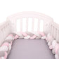 Un lit de bébé blanc avec un tour de lit tressé BABY PREMA Tour de Lit Tressé Noué aux couleurs pastel rose, blanc et gris tapissant l'intérieur. Le matelas a un drap-housse gris. Le berceau a des côtés à lattes et une tête de lit incurvée pour une protection supplémentaire.