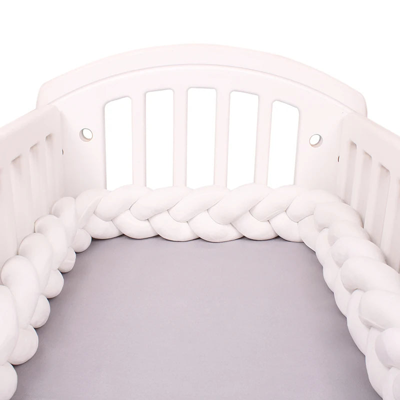 Un berceau blanc avec des côtés à lattes est présenté, contenant un tour de lit de protection tressé, ou Tour de Lit Tressé Noué de BABY PREMA, en blanc. Le matelas à l'intérieur du berceau est recouvert d'un drap gris clair. L'installation semble propre et confortable, conçue pour un sommeil sûr et confortable d'un bébé.