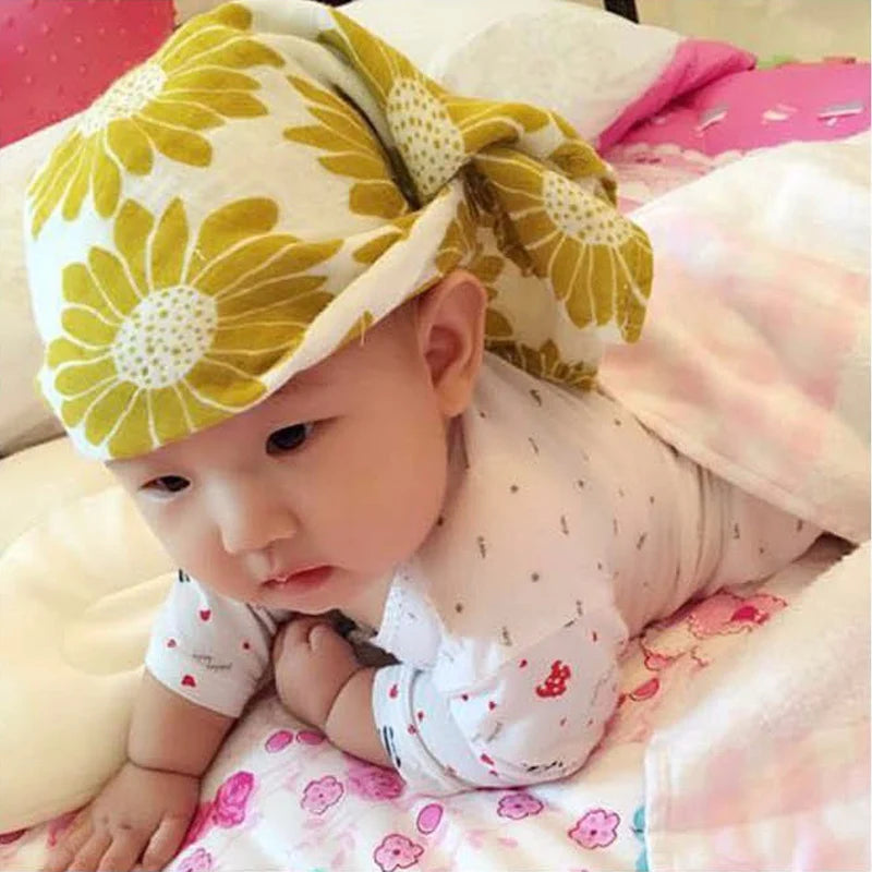 Un bébé avec une expression pensante portant un chapeau à motifs surdimensionnés reposant sur une couverture florale rose BABY PREMA en Lange Bébé en Coton et Fibre de Bambou.