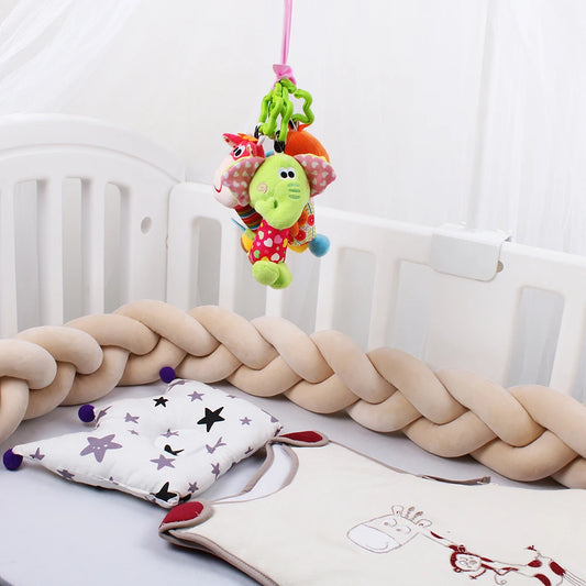 On voit un berceau confortable avec des tours de lit tressés Tour de lit bébé 4M de couleur claire. Au-dessus, un mobile suspendu présente des jouets en peluche colorés, dont un dinosaure vert. À l'intérieur du berceau, il y a un oreiller à motif étoilé et une couverture de couleur claire avec broderie girafe de BABY PREMA.