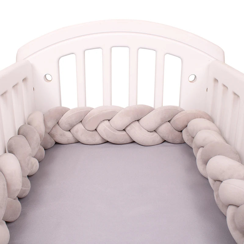 Un lit bébé blanc avec un Tour de Lit Tressé Noué gris tressé tapissant les côtés intérieurs. Le matelas est recouvert d'un drap doux gris clair, créant un environnement de sommeil sûr et confortable pour un bébé.