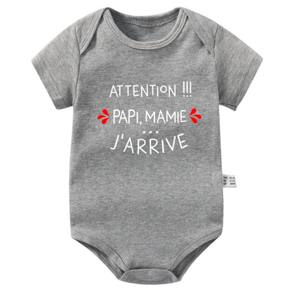 Un Body Manches Courtes Coton BABY-PREMA gris à manches courtes arbore le texte français "Attention !!! Papi, Mamie... J'arrive" en lettres blanches, orné de petits coeurs rouges de part et d'autre de "Attention !!! ". Cette barboteuse bébé à manches courtes en coton a une encolure ronde et une fermeture pressionnée dans le bas.