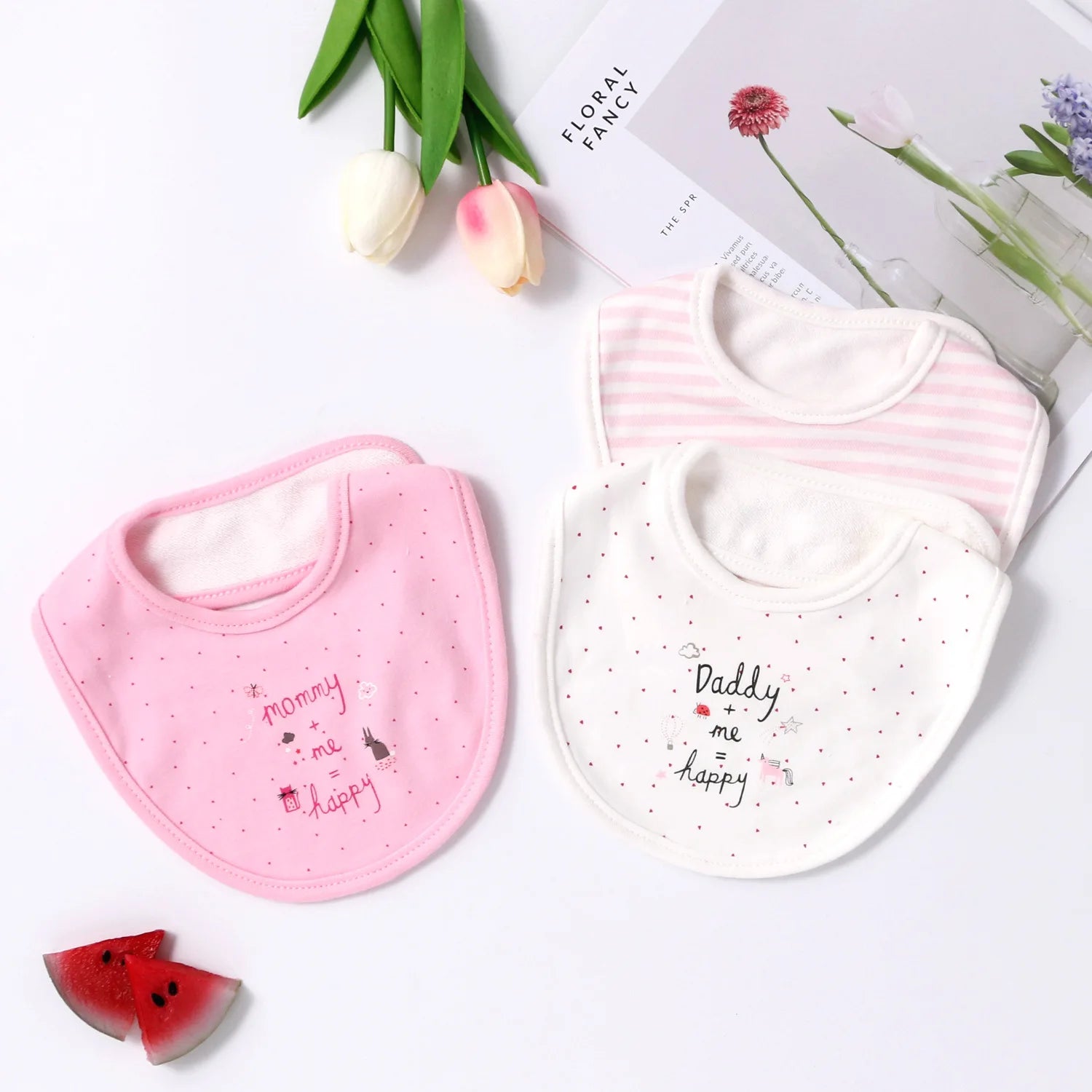 Trois Lot de 3 pièces Bavoirs Bébé de BABY-PREMA sont disposés sur une surface blanche. Deux sont blanches à petits pois et une est rose à petits pois. Un bavoir blanc indique « Papa me rend heureux » et le rose indique « Maman me rend heureuse ». Des tulipes et un livre floral sont en arrière-plan.