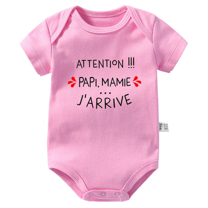 Un Body Manches Courtes Coton rose de BABY-PREMA avec des manches courtes comportant la phrase "Attention !!! Papi, Mamie... J'arrive" en texte noir avec des coeurs rouges de chaque côté. Ce body annonce nouveau-né comprend des fermetures à pression pratiques en bas.