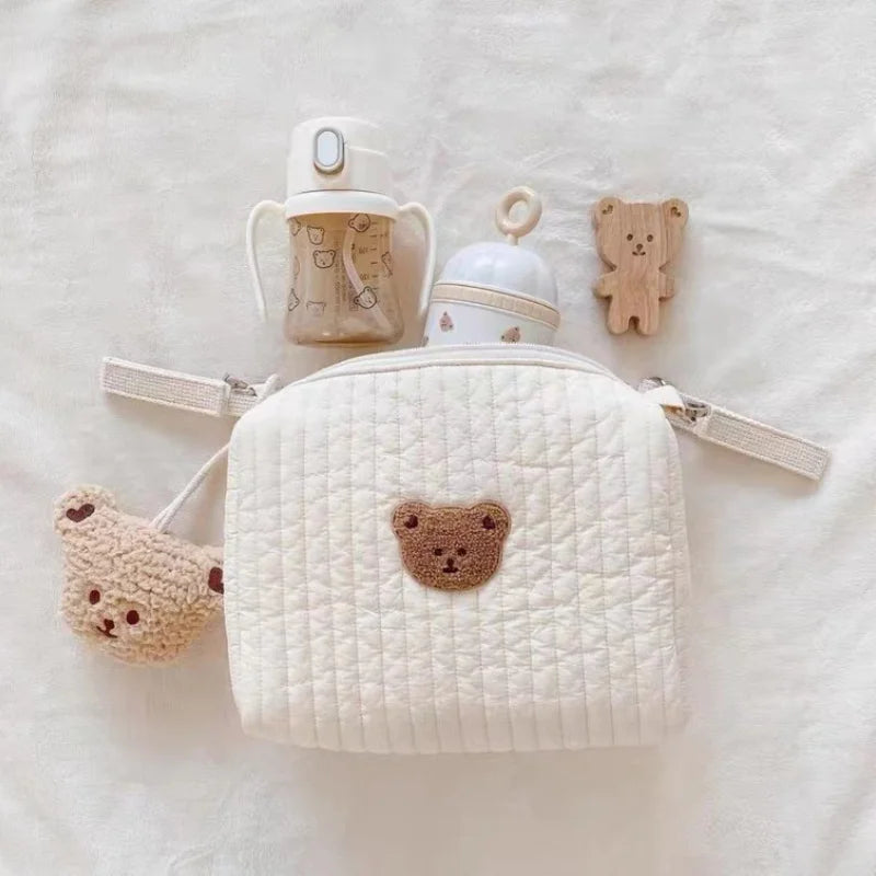 Une collection de BABY PREMA Sac de Couches Rangement Bébé soigneusement disposés sur un fond doux, comprenant un joli sac matelassé sur le thème de l'ours, un anneau de dentition en forme d'ours en peluche, un biberon à motifs et un gobelet assorti.