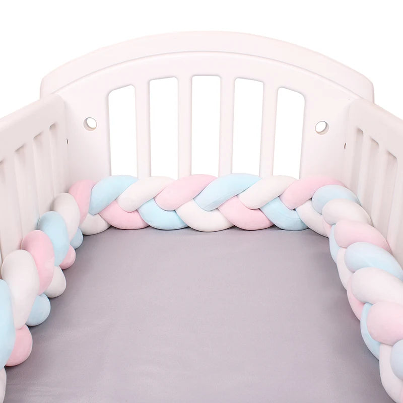 Un lit de bébé blanc avec un Tour de lit bébé 4M tressé de BABY PREMA avec un tour de lit tressé doublé à l'intérieur. Le pare-chocs de lit est fait de tissu doux et moelleux aux couleurs pastel, notamment des nuances de rose, de bleu et de blanc. Le berceau a des côtés à lattes et un surmatelas gris.