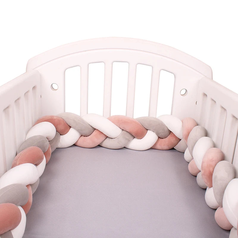 Un lit bébé blanc à lattes verticales et un Tour de lit bébé 4M tressé de BABY PREMA tapissant l'intérieur. Le pare-chocs présente un design doux et moelleux dans des couleurs rose pastel, blanc et gris, ressemblant à des oreillers tressés. Le matelas du berceau est recouvert d'un drap gris.