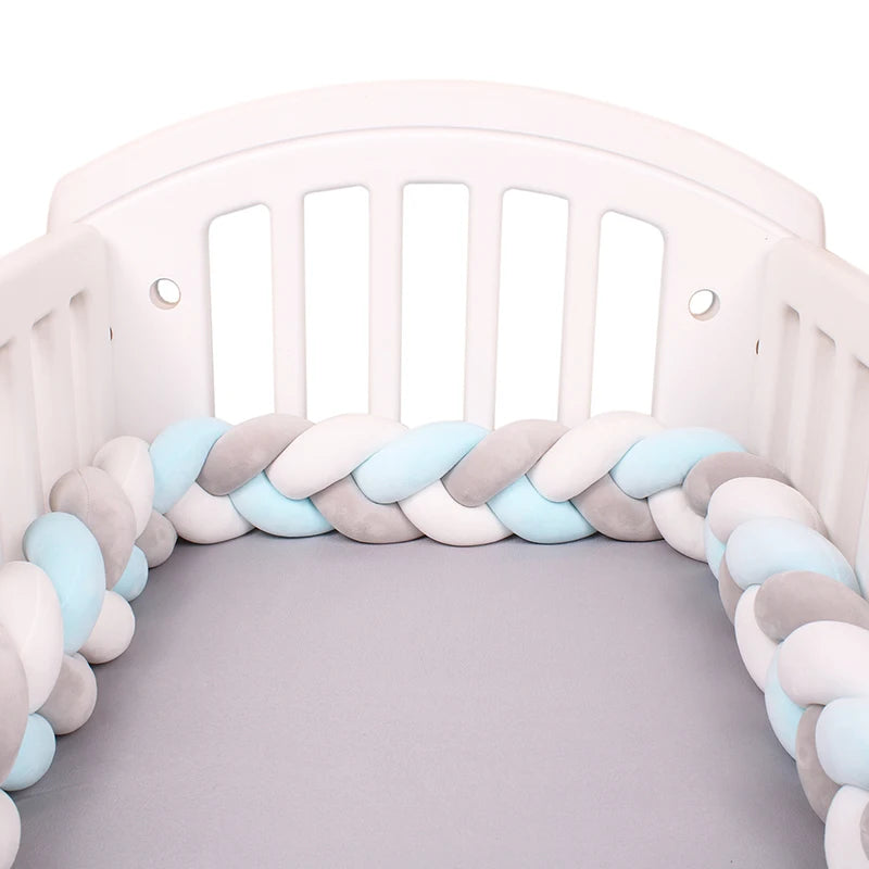 Un lit de bébé blanc comprend un Tour de lit bébé 4M tressé de BABY PREMA avec des pare-chocs tressés en tissu doux bleu, gris et blanc. Le berceau a des lattes blanches et le matelas est recouvert d'un drap uni gris. Le tour de lit tressé forme une barrière protectrice autour des bords du berceau.