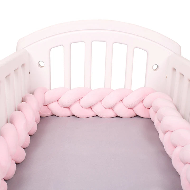 Un lit de bébé blanc avec un BABY PREMA Tour de Lit Tressé Noué rose clair bordant le périmètre intérieur. Le matelas du berceau est recouvert d'un drap gris clair. Le berceau a des lattes verticales sur les côtés et à l'arrière.