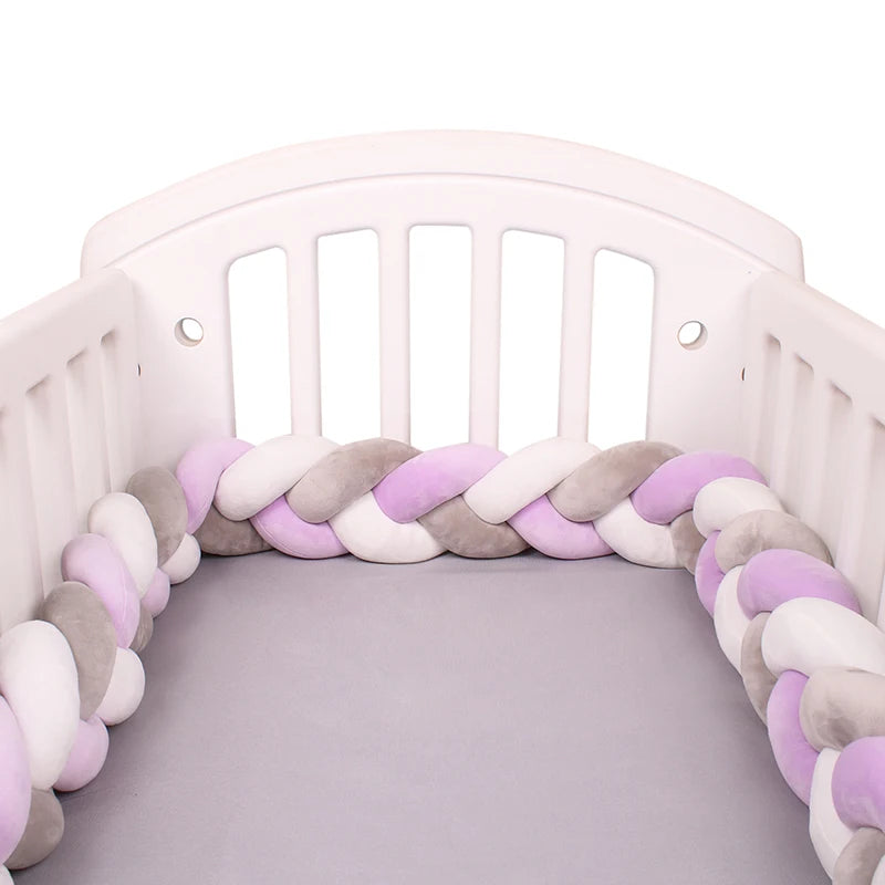 Un berceau blanc avec un Tour de Lit Tressé Noué de BABY PREMA, doté d'un tour de lit pastel tressé dans des tons de violet, rose, blanc et gris entourant les bords intérieurs. Le matelas est recouvert d'un drap uni gris. Le berceau a des lattes sur les côtés et à l'arrière.