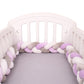 Un berceau blanc à lattes verticales contient un magnifique Tour de lit bébé 4M tressé de BABY PREMA avec des brins entrelacés en violet, gris et blanc. Le matelas est recouvert d'un drap gris clair.