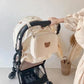 Main d'une personne ajustant un élégant sac à couches BABY PREMA Sac de Couches Rangement Bébé de couleur crème avec un joli motif d'ours sur une poussette de bébé, sur un fond minimaliste.