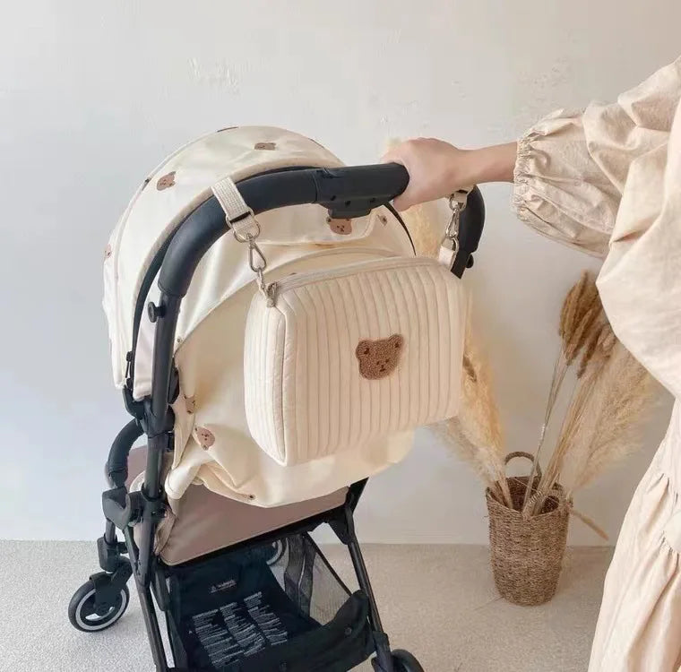 Main d'une personne ajustant un élégant sac à couches BABY PREMA Sac de Couches Rangement Bébé de couleur crème avec un joli motif d'ours sur une poussette de bébé, sur un fond minimaliste.