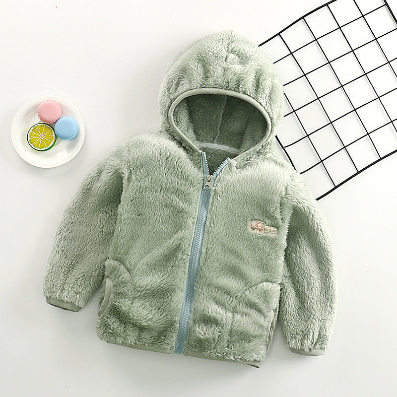 Un manteau Gilet pour bébé BABY PREMA vert pastel doux posé à plat à côté d'une assiette avec des macarons colorés et un fond à motifs géométriques noir et blanc.