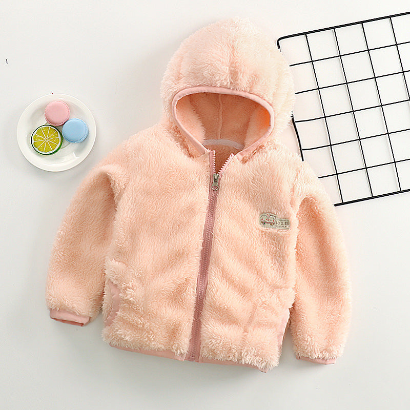 Une veste polaire à capuche BABY PREMA prématuré rose douillette, exposée à côté d'une assiette décorative avec des macarons colorés et un carnet à motifs géométriques.