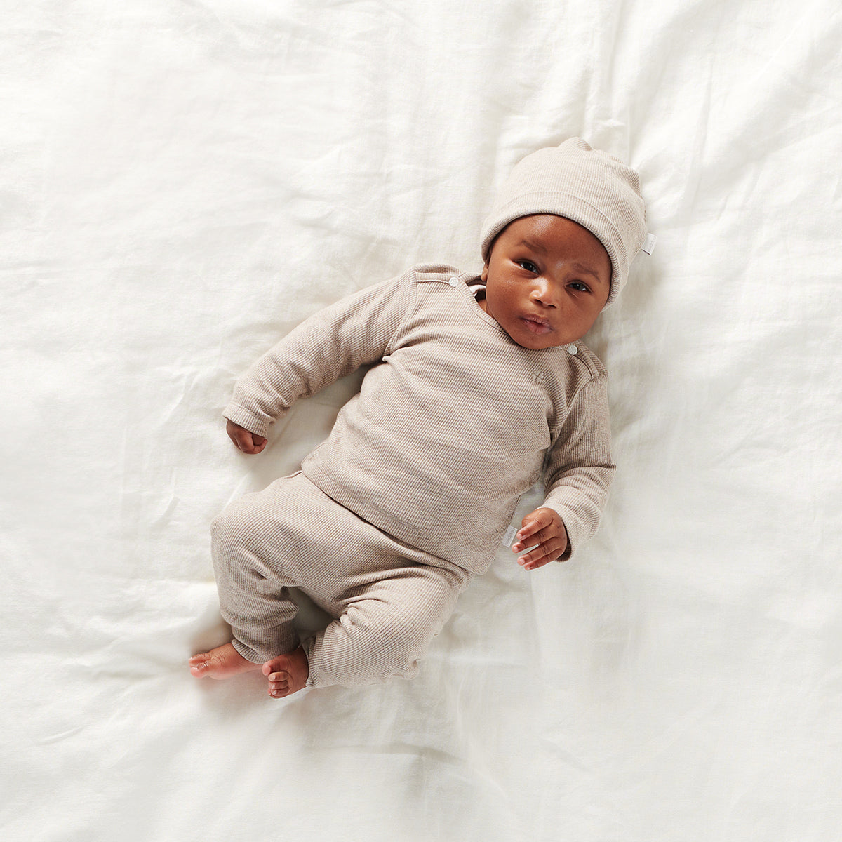 Un bébé serein vêtu d'une tenue beige douillette et d'un bonnet Bonnets tricot Blanc assorti repose confortablement sur une douce couverture blanche, entouré des accessoires d'hygiène nécessaires pour bébés de NOPPIES.