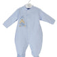Une combinaison bébé bleu clair à manches longues et bas à pattes, ornée d'un ours brodé avec une couronne sur la poitrine, accrochée à un cintre en plastique - le complément parfait à tout trousseau de naissance. Il s'agit du Pyjama Blanc en Coton - King Bear de DANDELION.