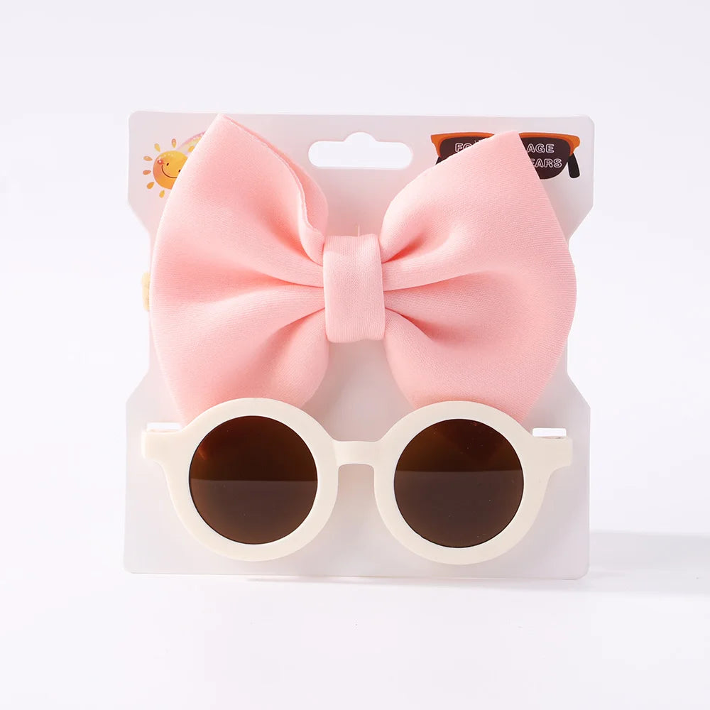 Un ensemble d'accessoires pour enfants, comprenant un grand nœud rose, des bandeaux à nœuds en nylon et des lunettes de soleil pour bébé rondes blanches avec des verres foncés est l'Ensemble d'Accessoires Bébé | Lunettes de Cheveux pour Bébé de BABY PREMA.