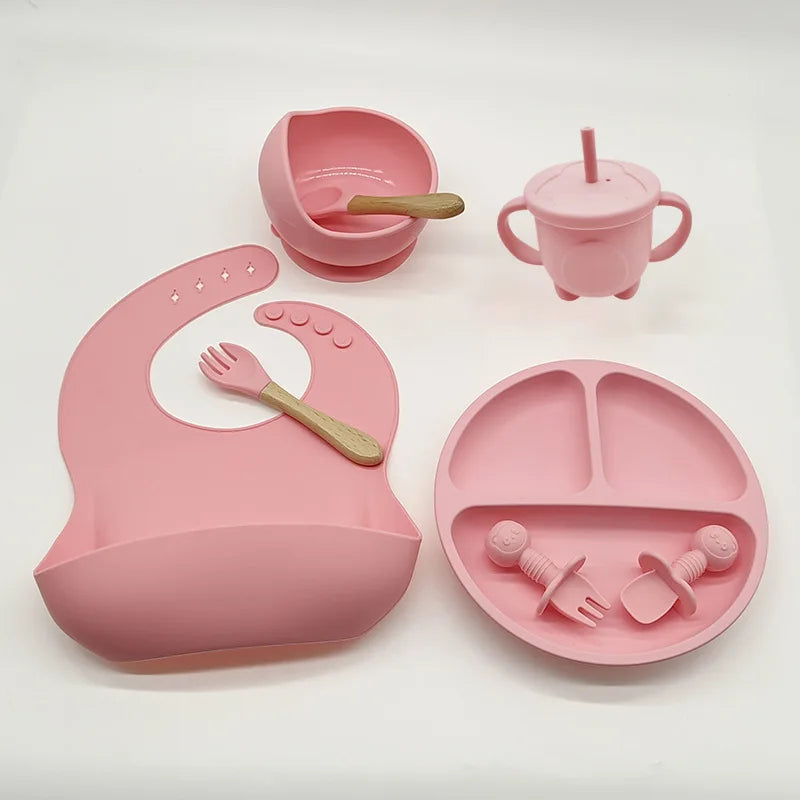 Un ensemble d'articles d'allaitement roses en silicone de haute qualité comprenant une assiette, un bavoir, une tasse avec poignées, deux bols à ventouse, une cuillère et deux ustensiles en forme de fourchette et de cuillère a été remplacé par l'Ensemble 6-8. Pièces de Vaisselle pour enfants par BABY PREMA.