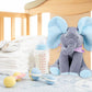 Un Doudou Éléphant Musicale BABY-PREMA assis sur le lit d'un bébé prématuré, avec un biberon, une paire de petites chaussures blanches et deux maracas jouets à proximité, créant une chambre de bébé chaleureuse et nourrissante.