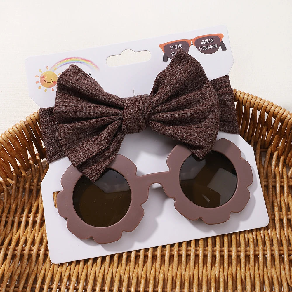 Un présentoir d'Ensemble d'Accessoires Bébé en forme de fleur marron | Lunettes de Cheveux pour Bébé de BABY PREMA posées sur une carte blanche, posée sur un panier en osier.