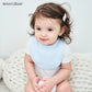 Un jeune enfant aux cheveux ébouriffés et au regard curieux portant un bavoir bleu conçu pour Bavoir Bébé en Coton Bio, confortablement assis sur une couverture tricotée BABY PREMA.