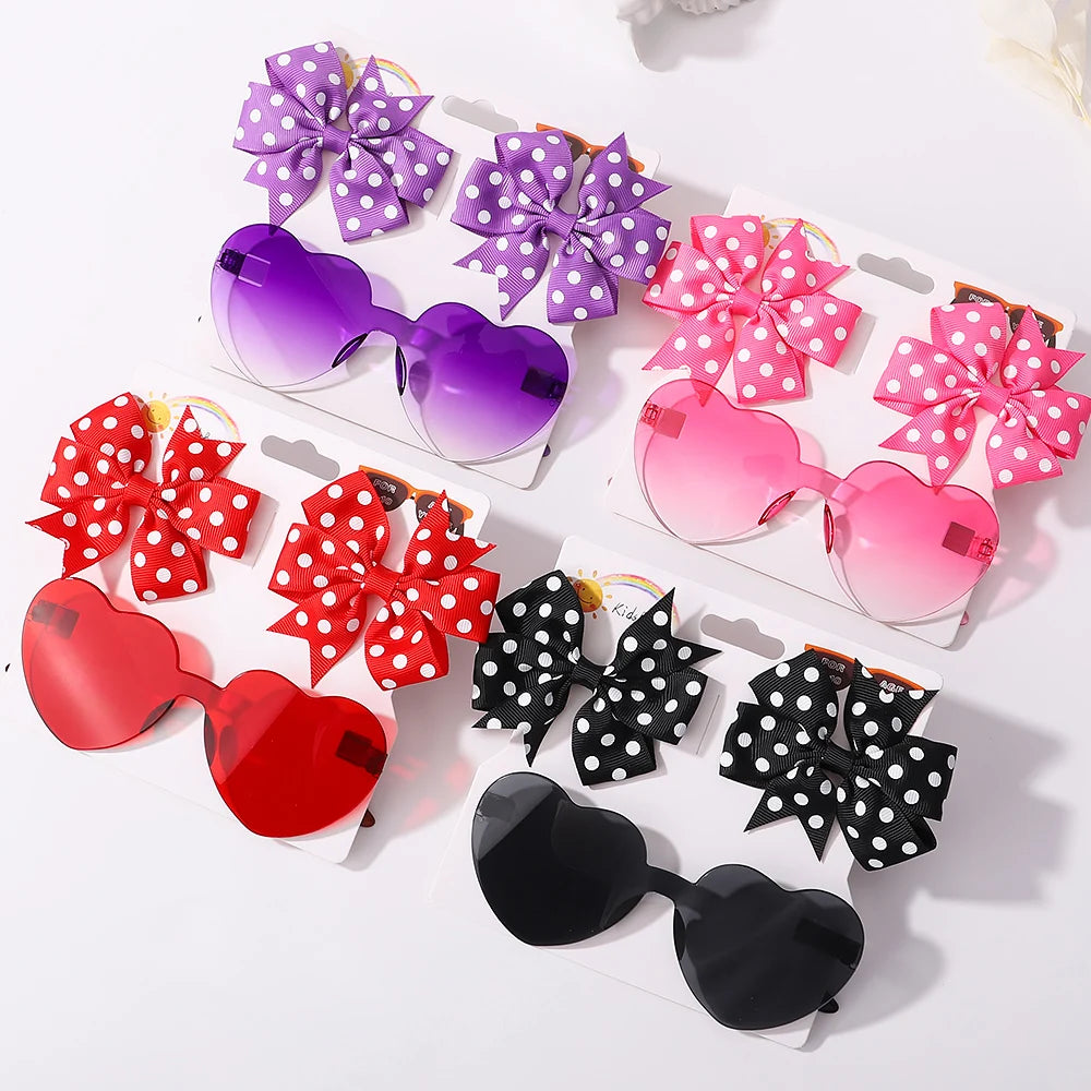 Quatre paires de lunettes de soleil pour bébé en forme de cœur sont chacune accompagnées d'un bandeau à nœud en nylon assorti en rouge, violet, rose et noir, le tout affiché sur un fond blanc. Présentation de l'ensemble d'accessoires bébé | Lunettes de Cheveux pour Bébé de BABY PREMA.