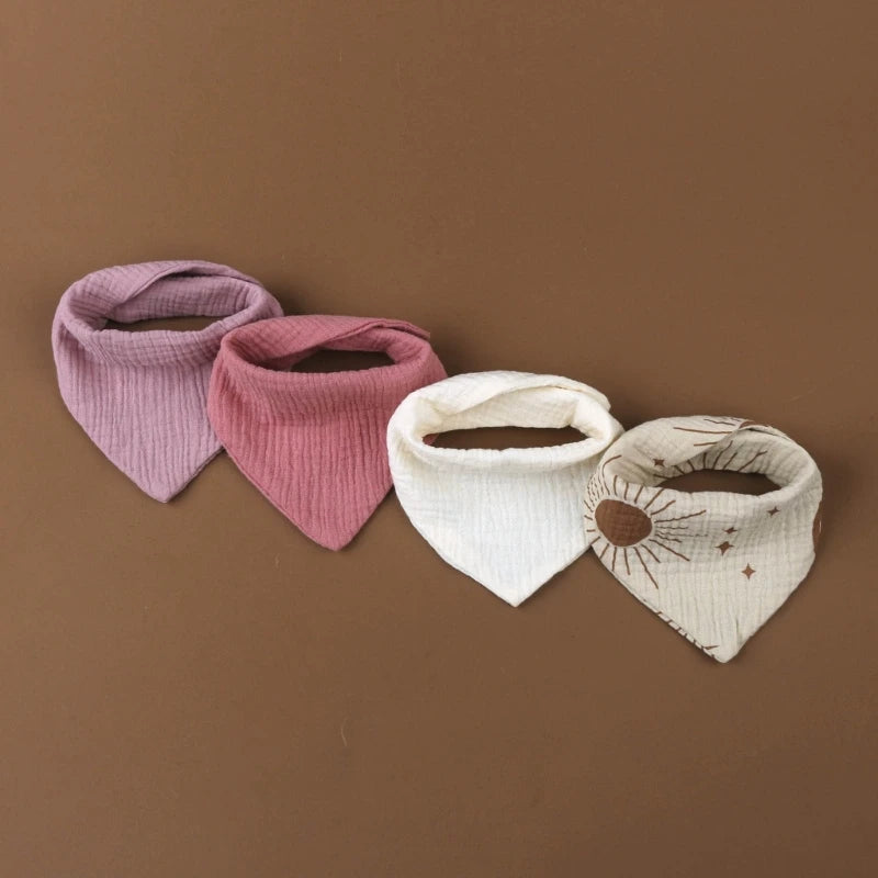 Quatre bavoirs bandana pour bébé sont soigneusement placés sur un fond marron. De gauche à droite, les couleurs sont lavande, marron, blanc et beige avec un motif soleil et étoile. Les Bavoirs en mousseline coton pour bébé de BABY-PREMA sont fabriqués dans un tissu doux et texturé et ont une forme triangulaire.